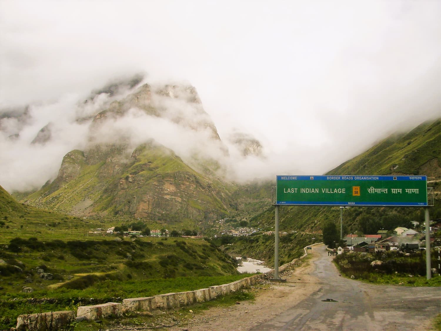 The road into Mana with a signboard announcing the last Indian village and green slopes behind